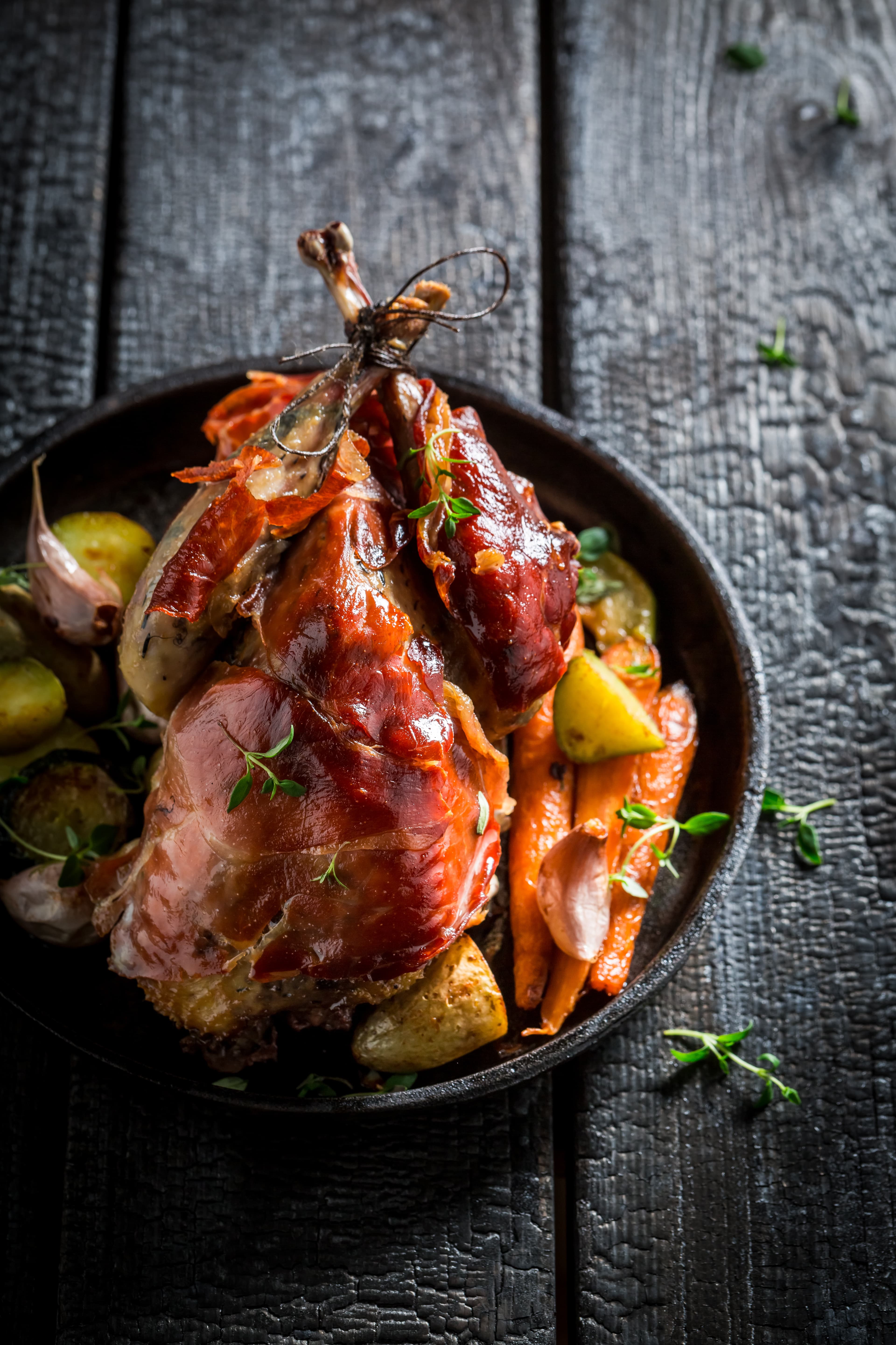 A whole pheasant wrapped in bacon, roasted with carrots and garlic in a dark pan.