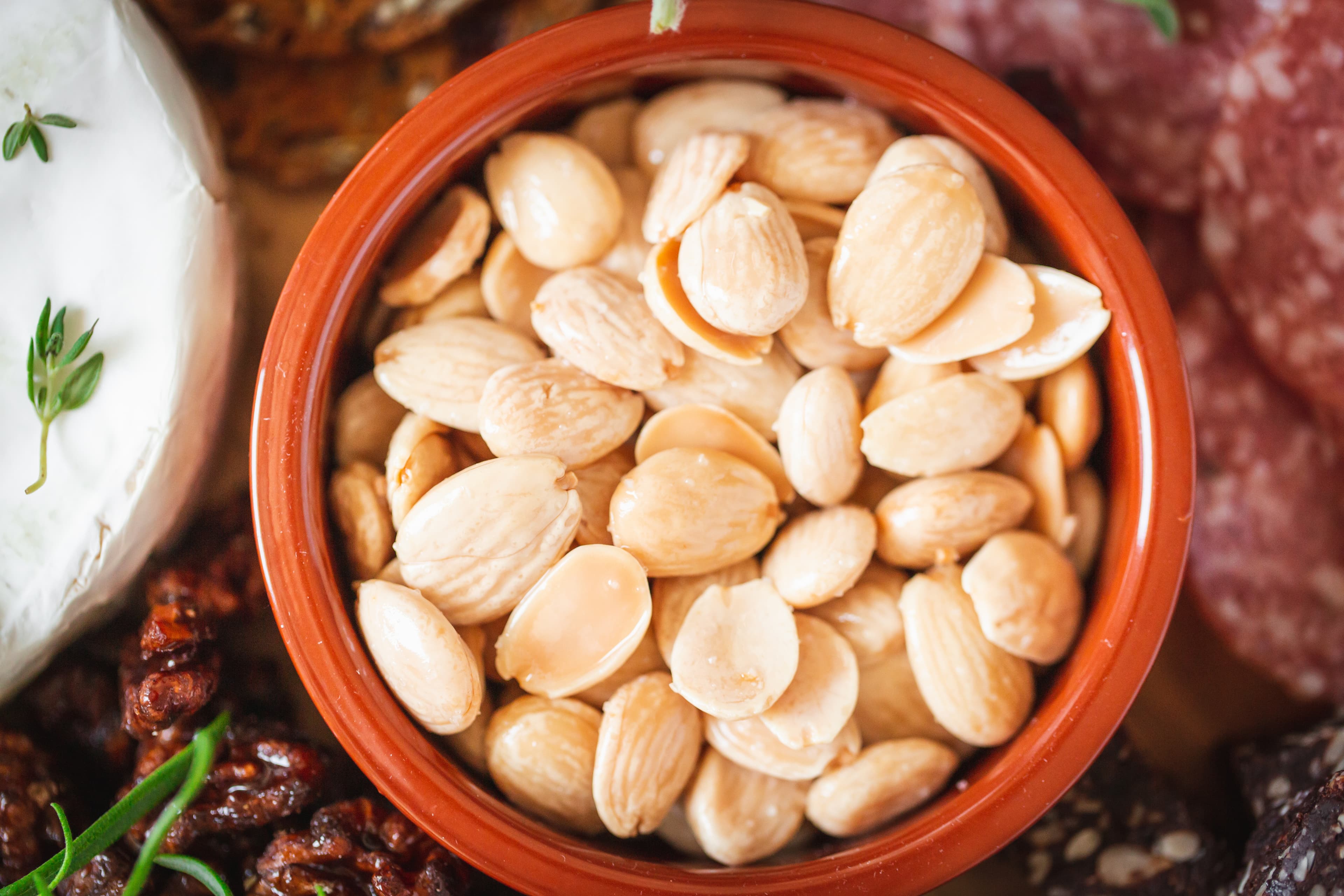 Blanched almonds in a terracotta bowl beside brie, walnuts, and sliced salami.