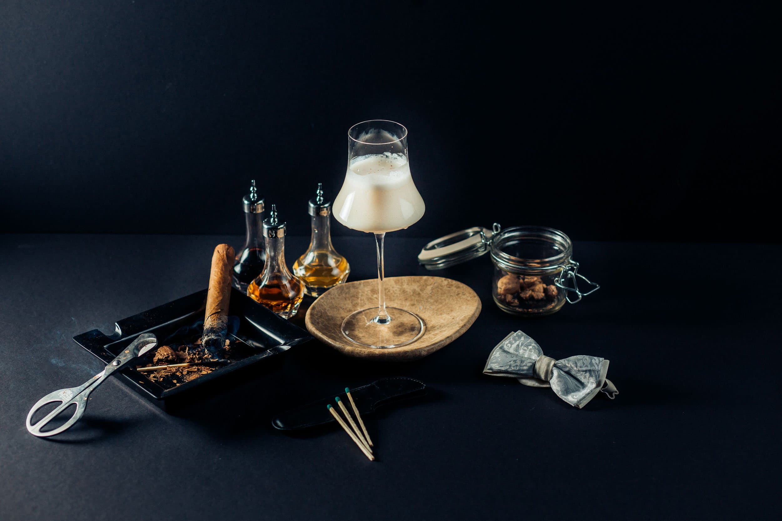 A creamy cocktail in a stemmed glass beside a lit cigar, matches, and small bottles.