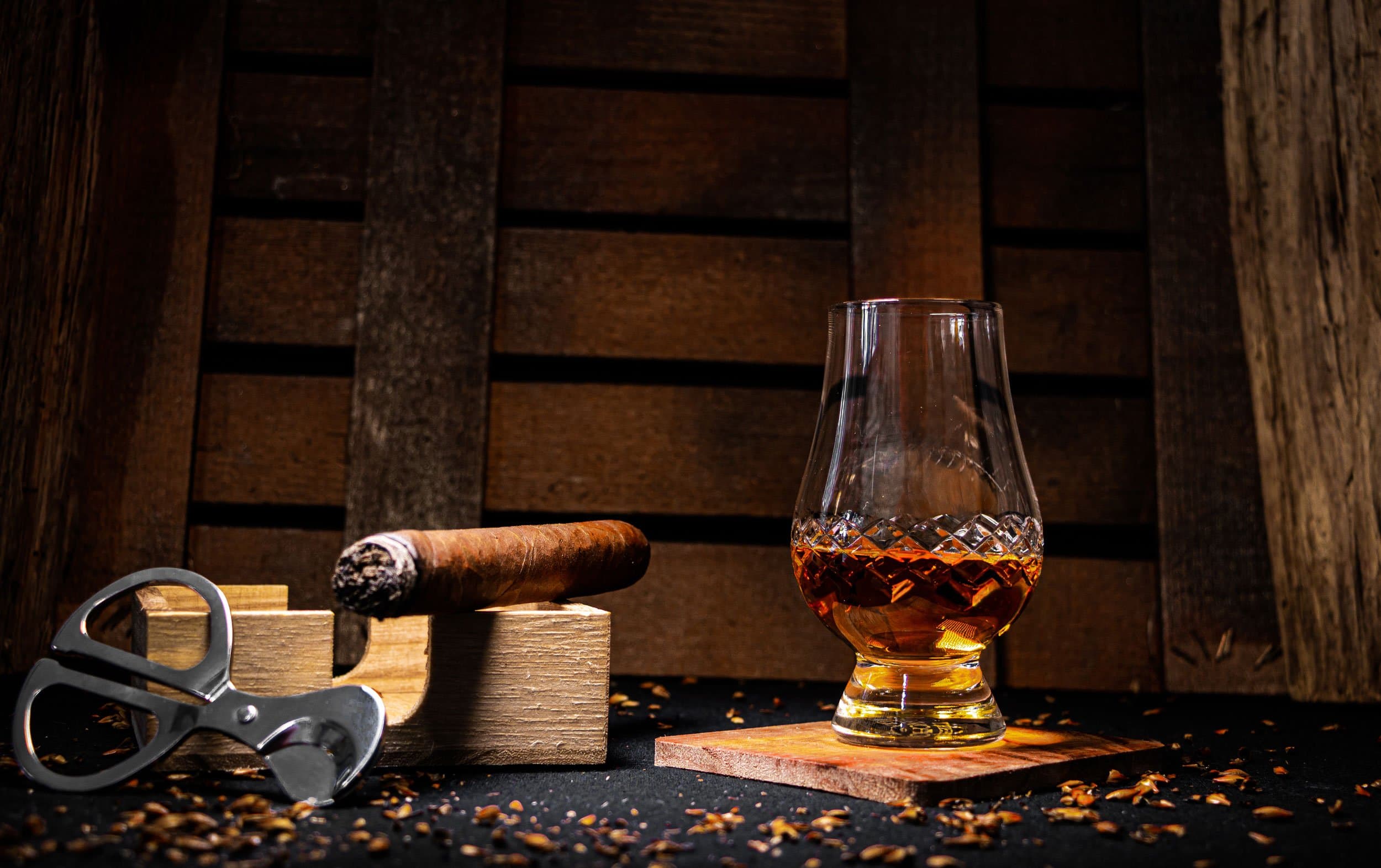 A tulip glass of amber spirit beside a cigar on a wooden rest and a cutter.