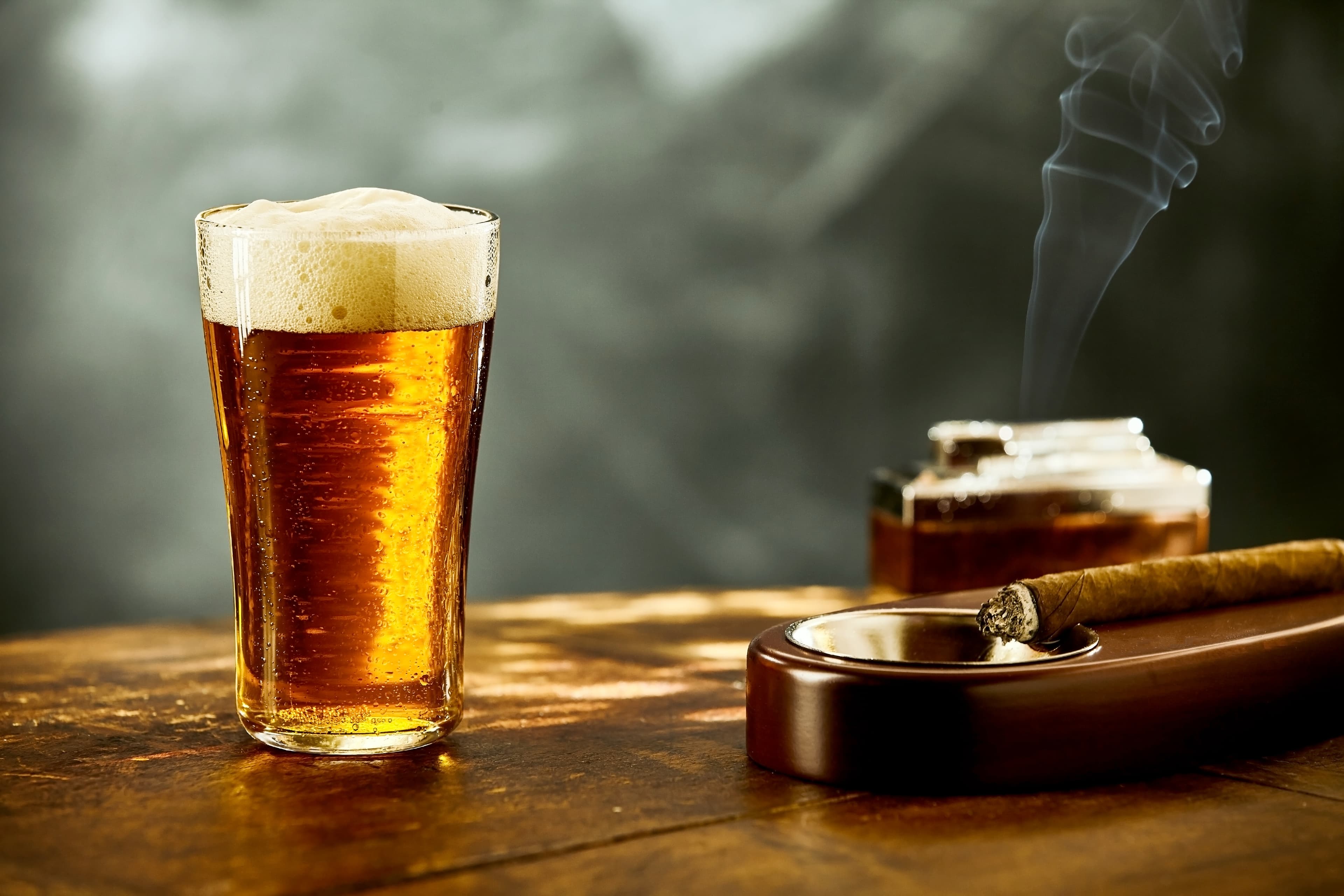 A pint of amber beer beside a lit cigar in a wooden ashtray.