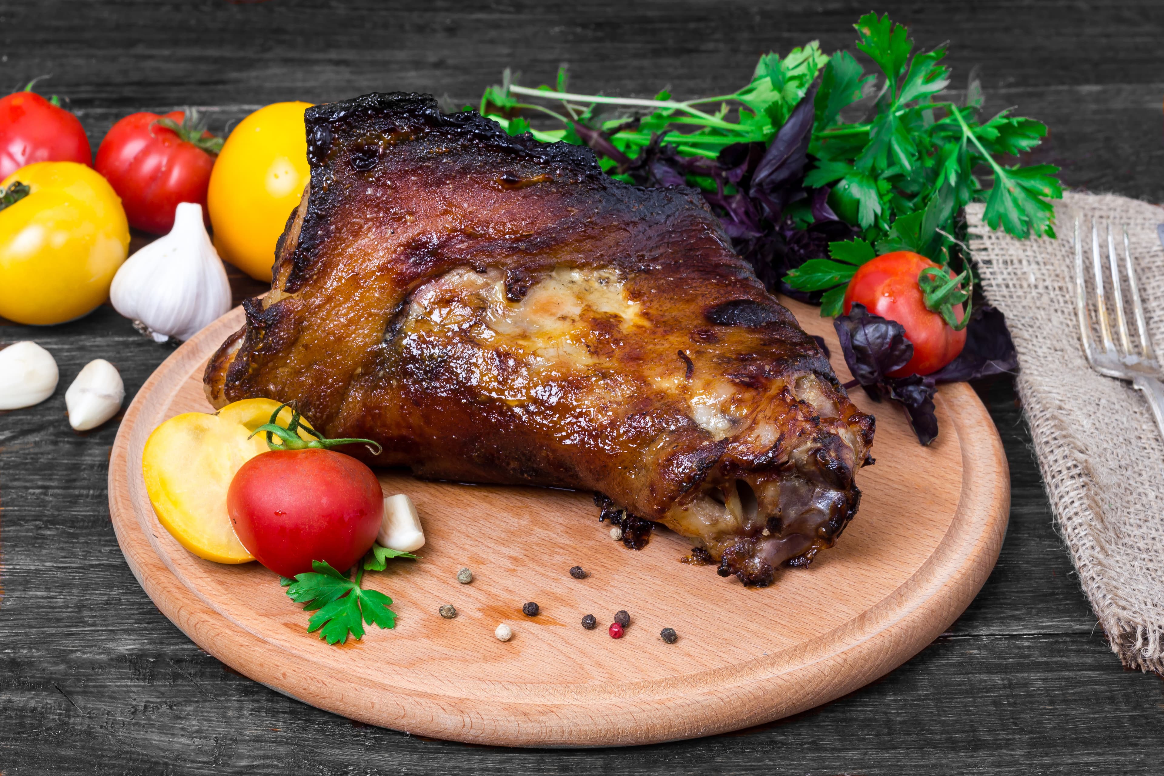A roasted pork knuckle with crackling, tomatoes, garlic, and herbs on a wooden board.