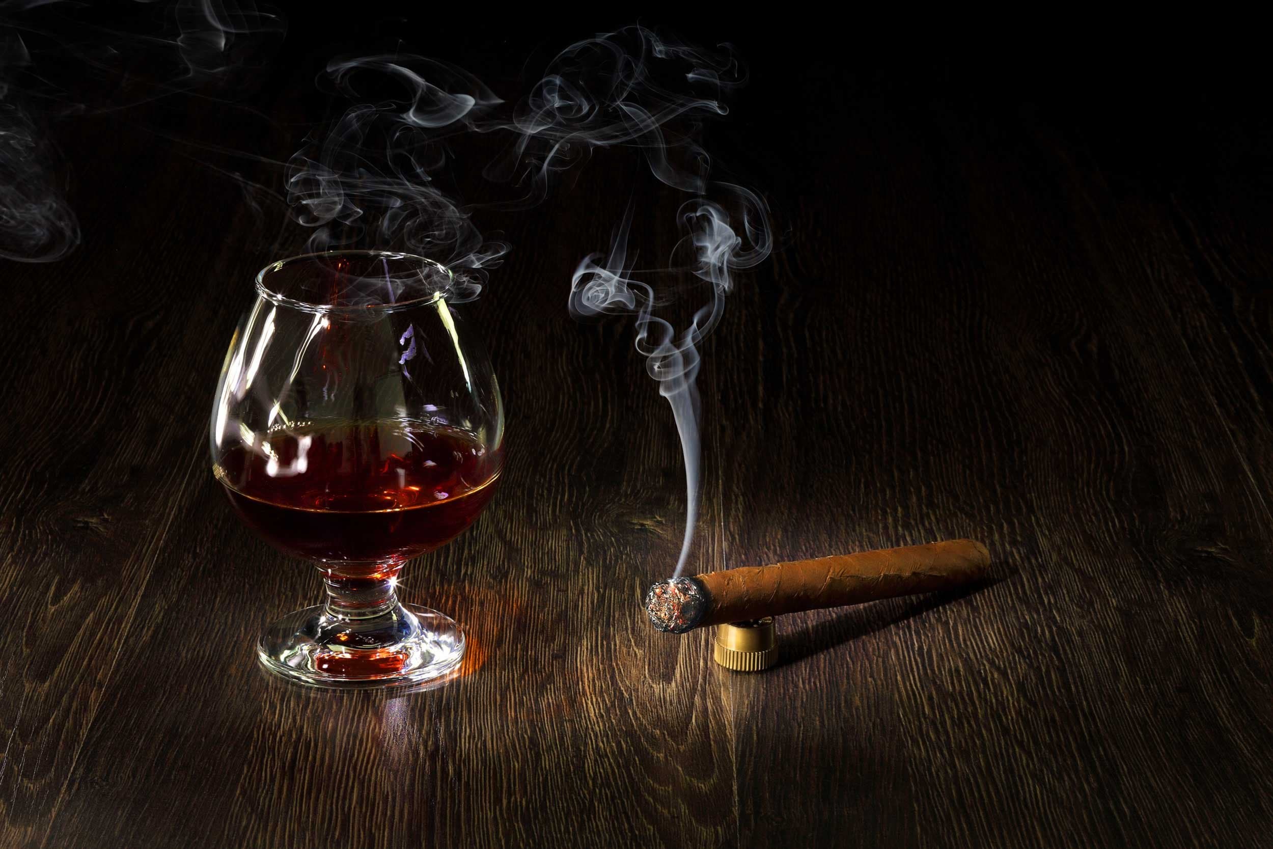 A lit cigar on a brass rest beside a snifter of amber spirit, smoke rising.
