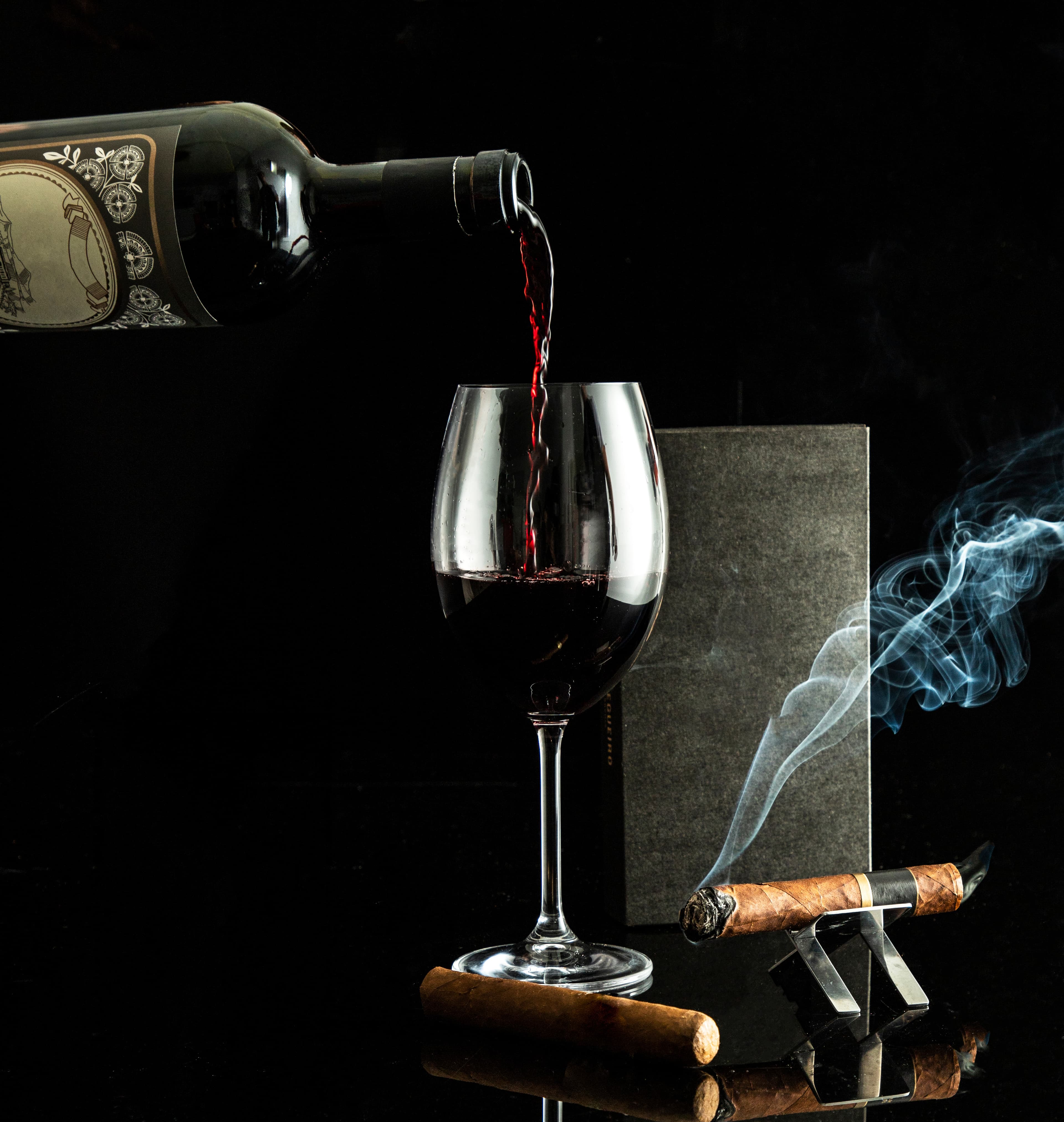 Red wine poured into a stemmed glass beside a lit cigar on a silver stand.